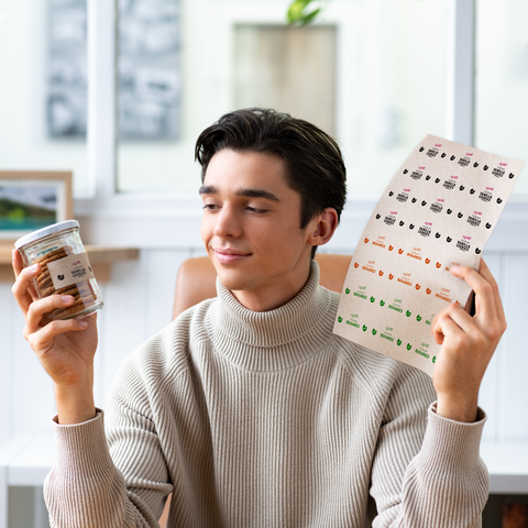 Ein junger Mann hält in der einen Hand ein Glas mit Snacks und in der anderen ein Etikett mit verschiedenen Designs.